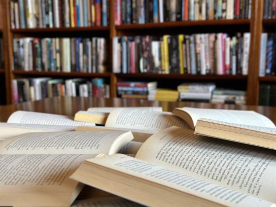 Stack of opened books on a desk with bookshelf in the background.